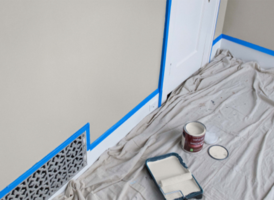 Drop cloth next to masked-off wall. Off-white paint and roller in tray. Open can of paint with lid off. 