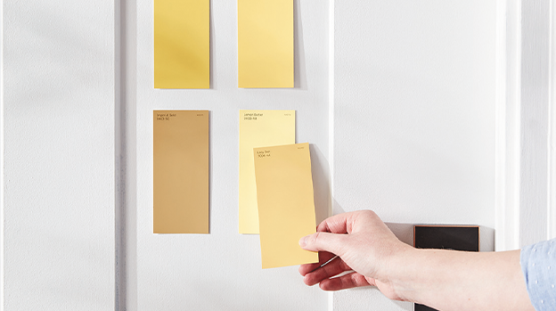 Person holding paint chips up to bathroom wall with sink, mirror and light fixture in background.