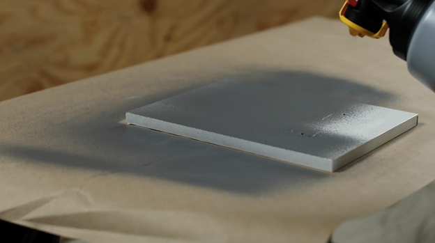 Sprayer spraying gray paint on white wood board.