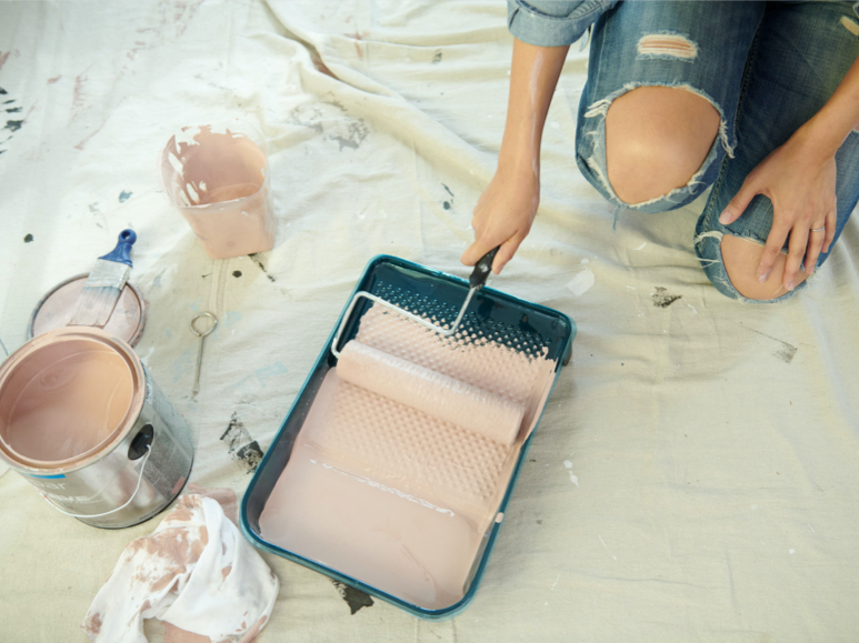 Person holding roller in tray on top of drop cloth with a can, brush and rag in foreground.